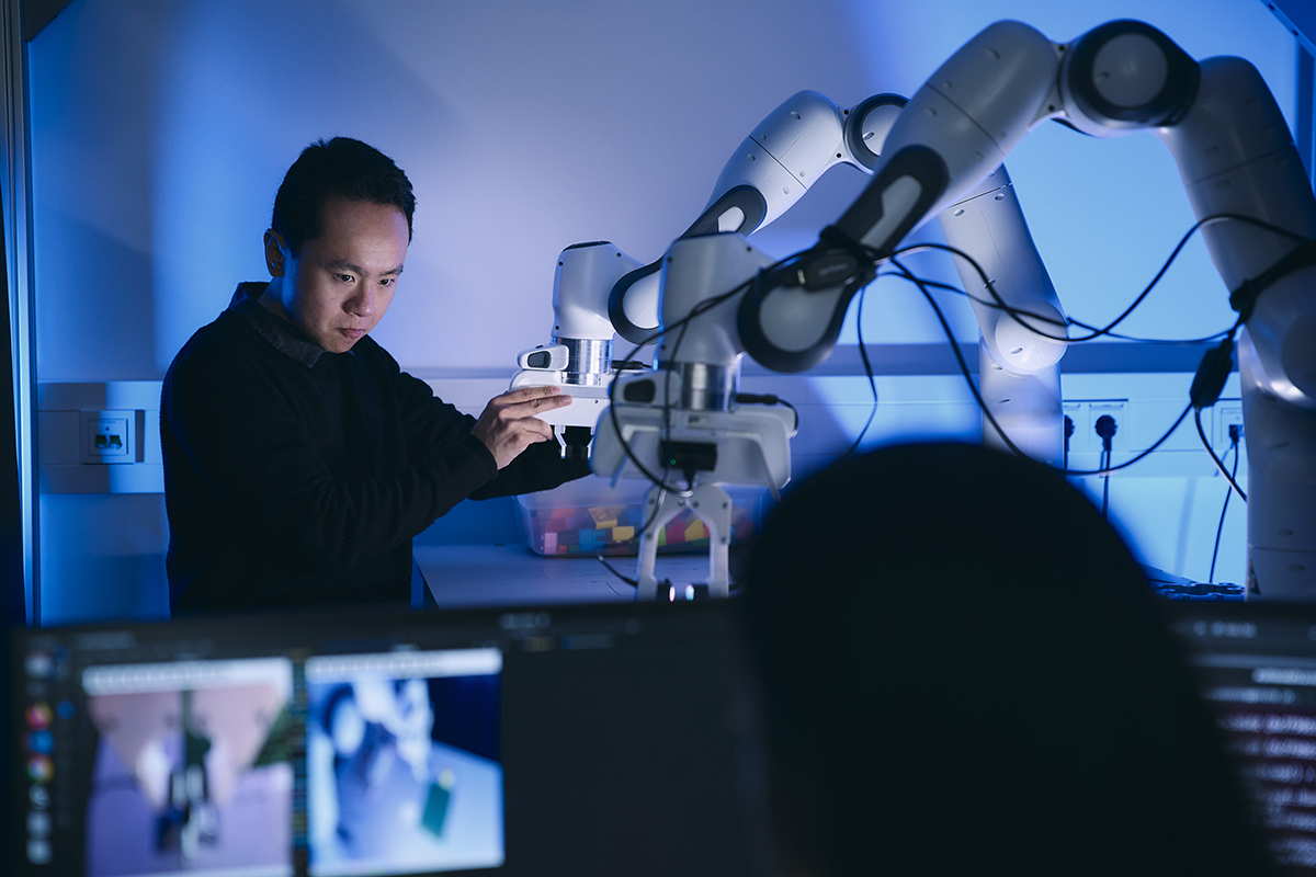 Mensch bedient Roboterarm im Labor, mehrere Roboterarme arbeiten an einem Tisch.