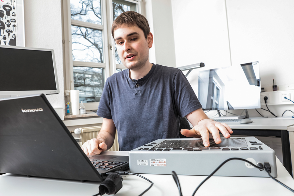 Junger Mann arbeitet an einem Laptop und einem Eingabegerät für sehbeeinträchtigte Studierende in einem  Büroraum.