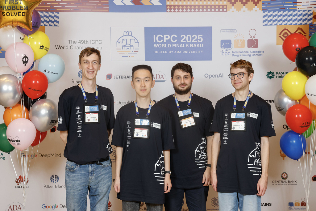 Vier Teilnehmer der ICPC 2025 World Finals mit Veranstaltungsbanner und bunten Luftballons im Hintergrund.