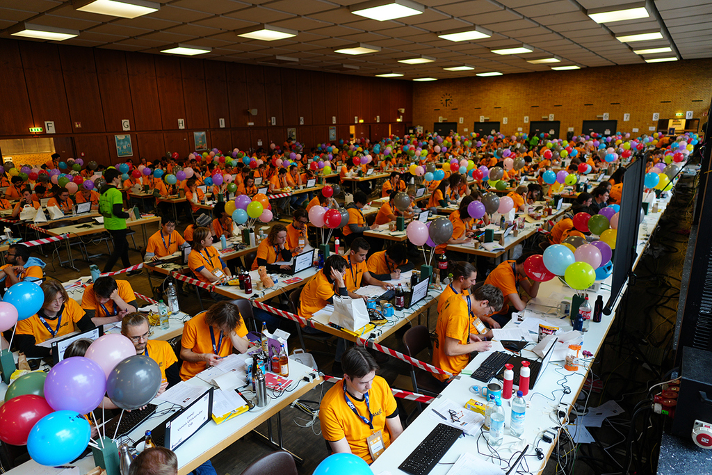 Großer Saal mit vielen Tischen, Teilnehmern in Orange, bunten Ballons und Laptops.